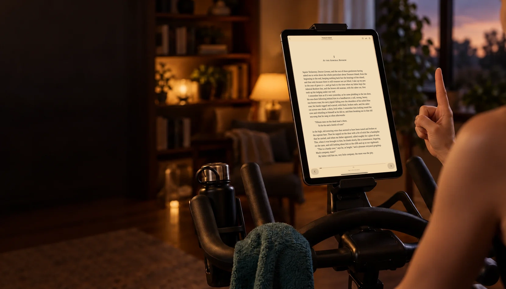 A tablet mounted on an exercise bike in a warmly lit room, showing the real PageFlick iPad reading view with a raised index finger beside it